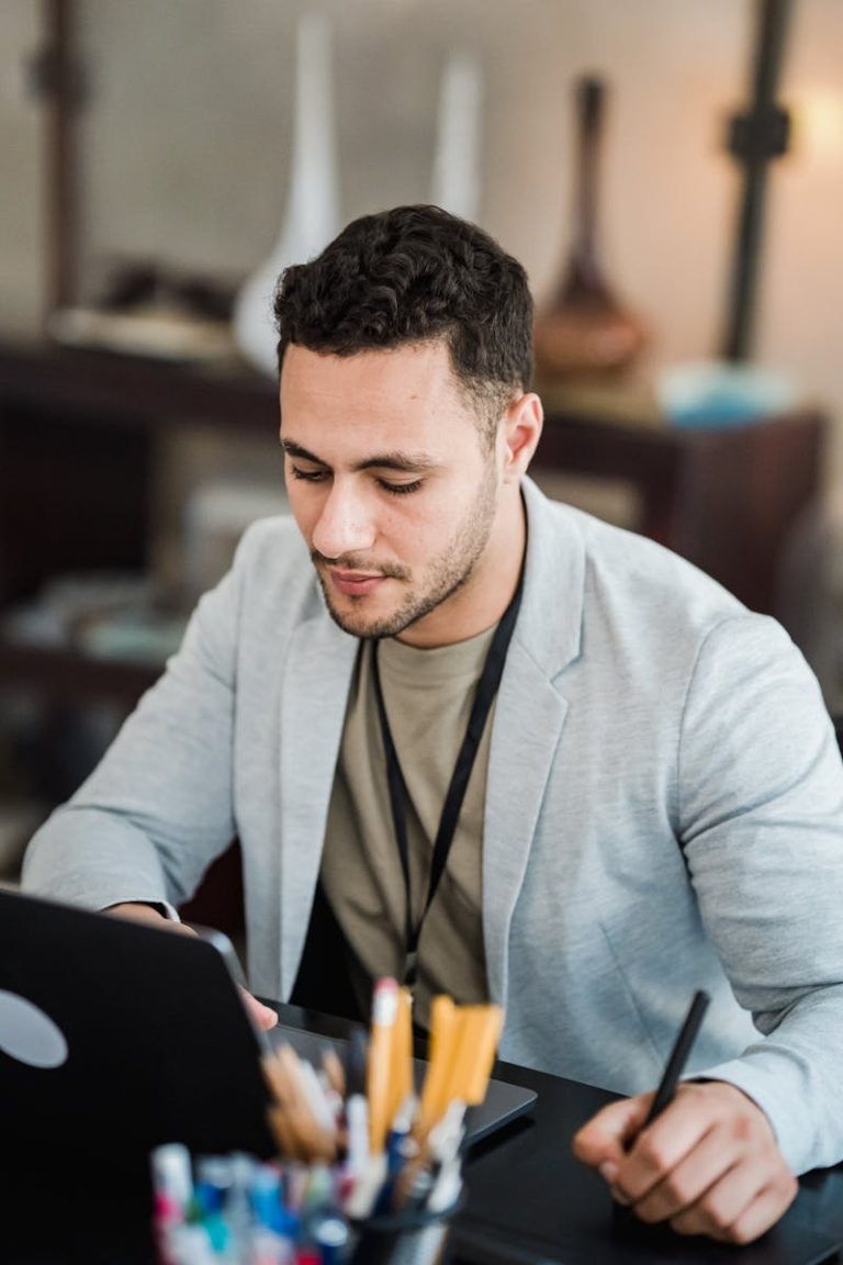 A young professional in a suit works intently on a laptop and tablet at an office desk.